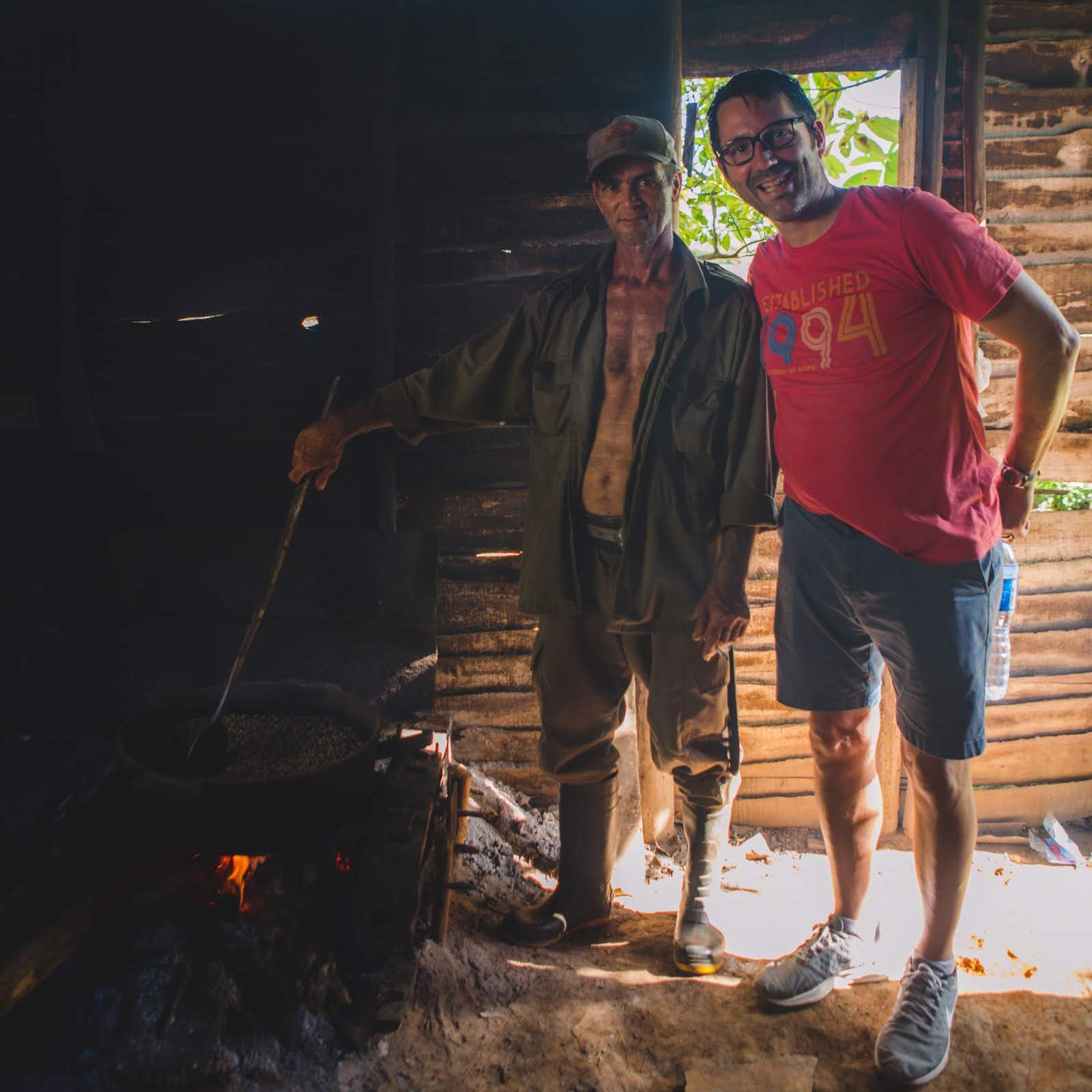 Gardner Tours Custom Cuba Tours Man poses with a Cuban Coffee Farmer as they roast coffee over and open fire in a hut