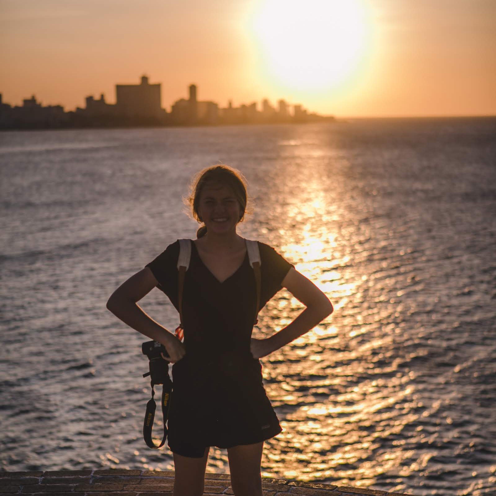 Gardner Tours Custom Cuba Tours Young Woman Holding her Camera as she stands in front of the sunset on El Morro Castle overlooking Havana