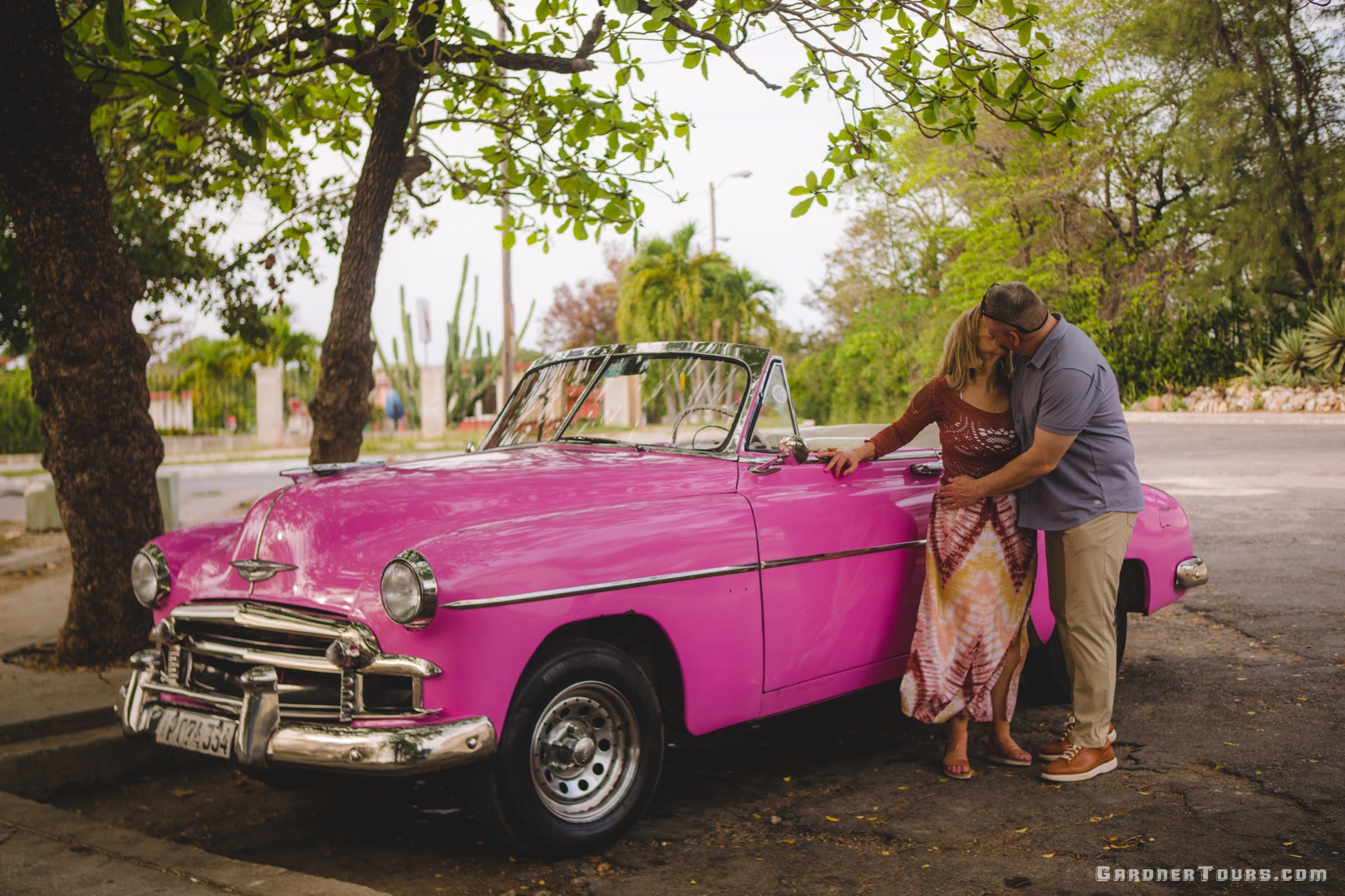 A middle aged white couple kiss in front of a pink 1950 Chevrolet convertible under the shade in Havana Cuba