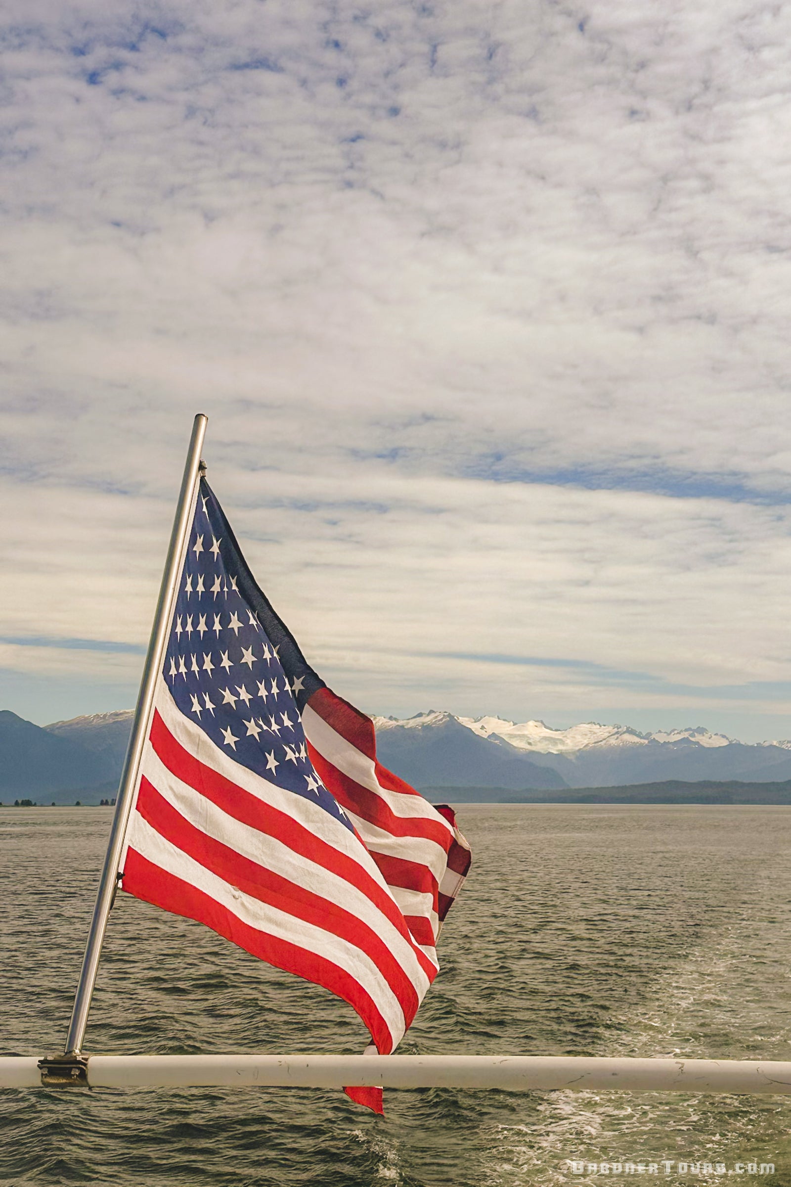 The American flag flying from a yacht with snow-capped Alaskan mountains and glaciers in the distance.