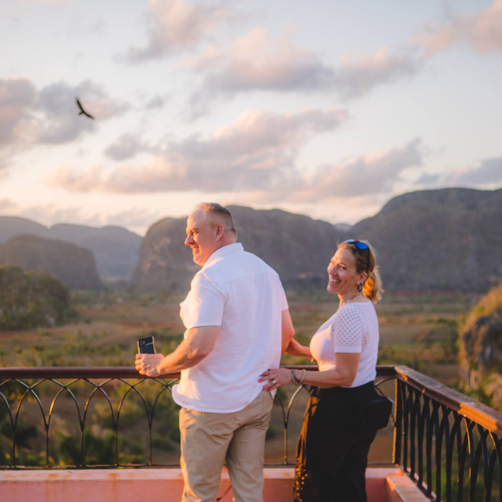 A sweet couple on their Luxury Cuba Tour, with Gardner Tours, wearing matching white outfits and laughing while enjoying the sunset at the Los Jazmines Viewpoint overlooking the Vinales Valley in Vinales Cuba.