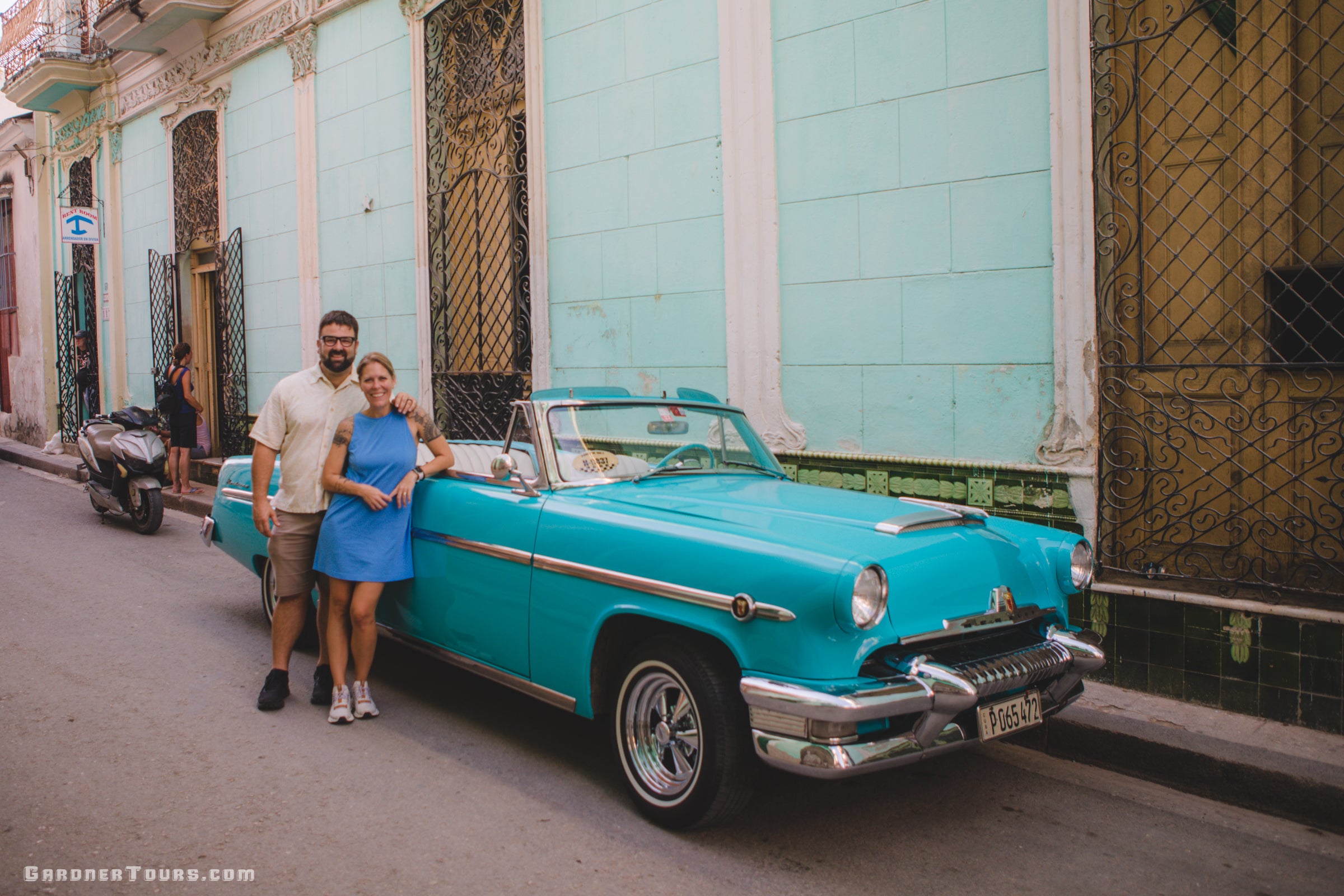 Two people standing next to a vintage turquoise car on a street in colorful Havana, Cuba.