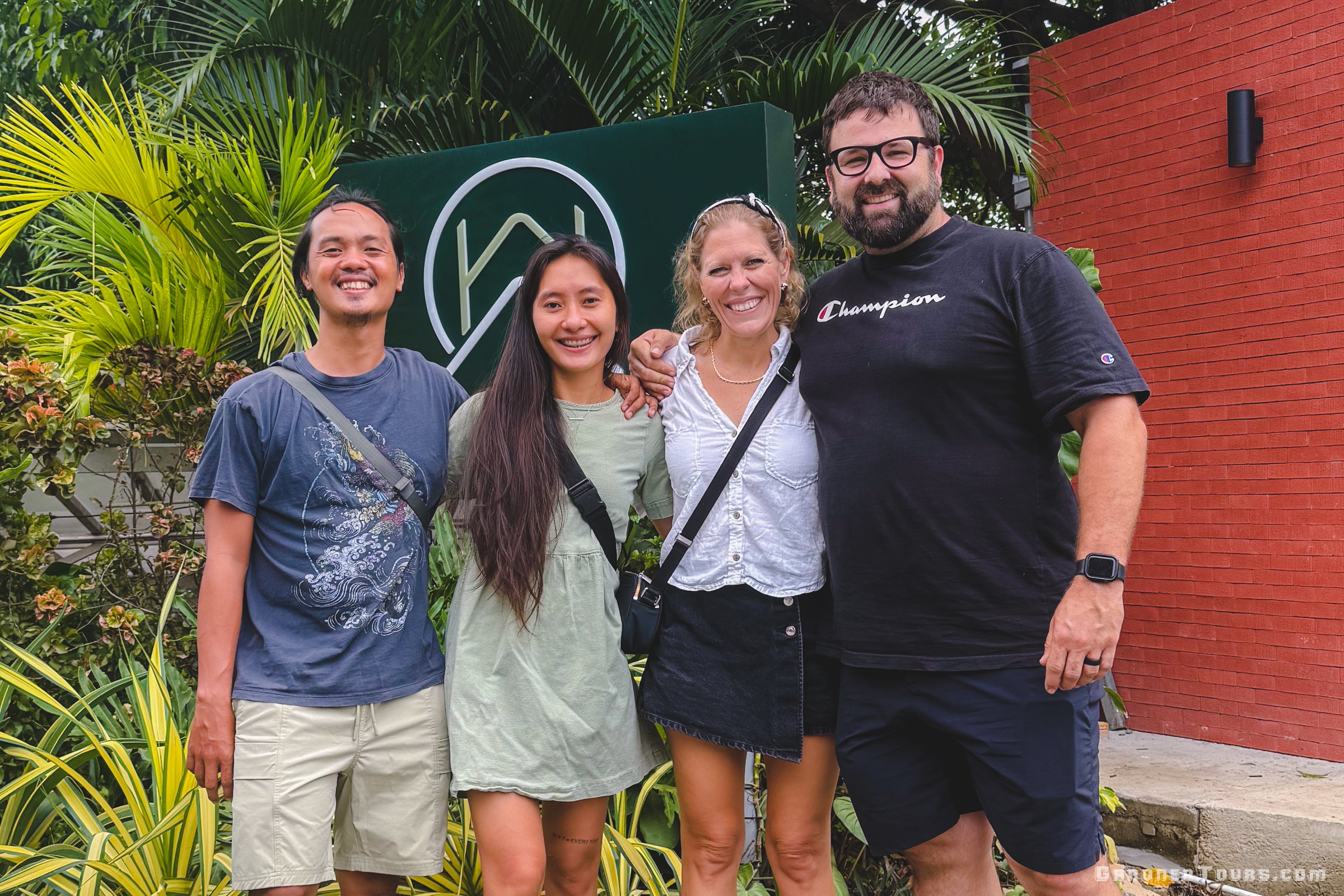 Four people posing together in front of a green sign with a logo, surrounded by plants.