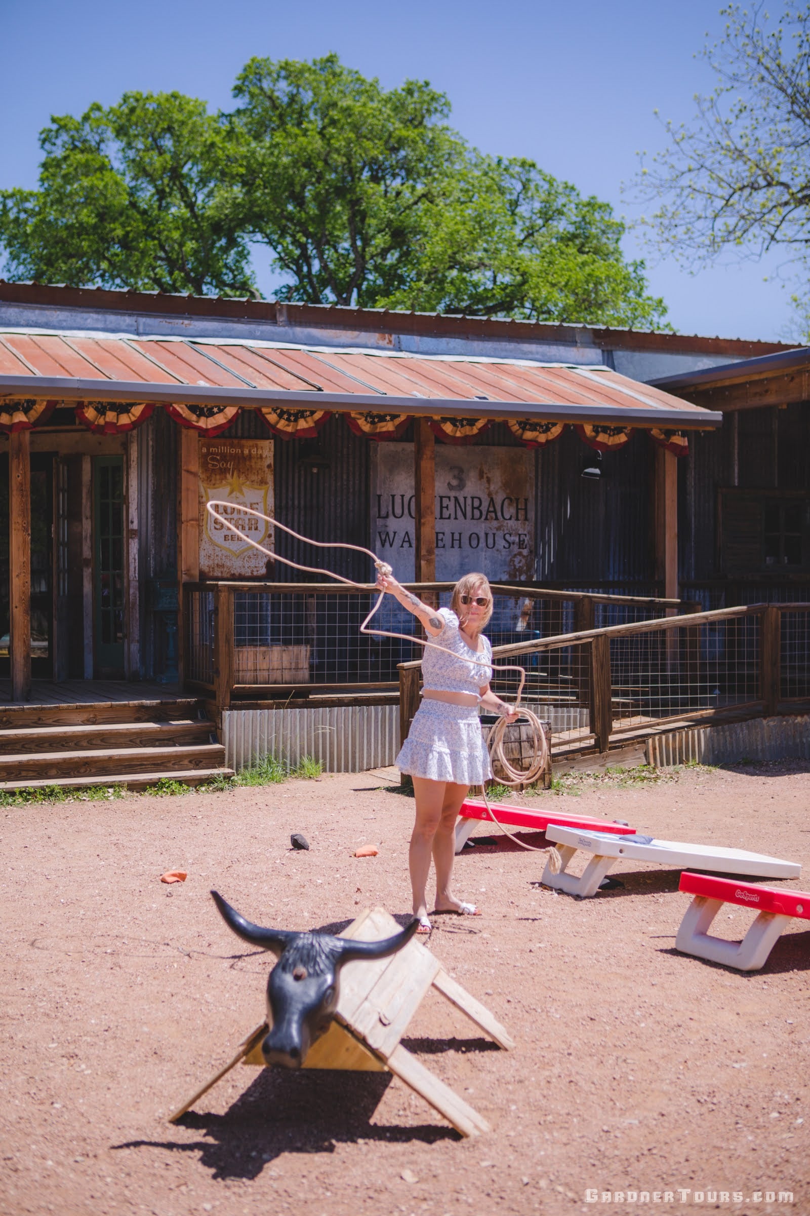 Woman holding a lasso in front of a wooden building with a sign, with a steer head and seesaw in the foreground.