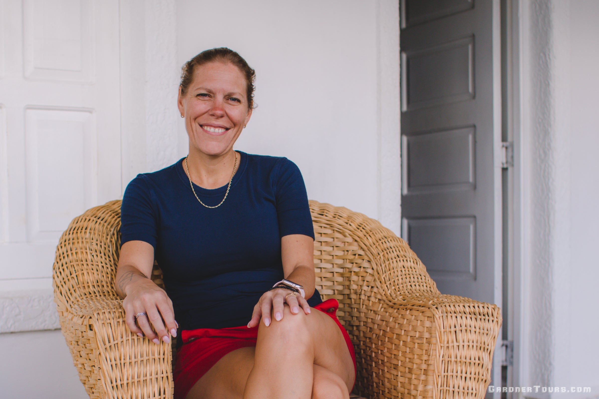 woman wearing a dark blue shirt and red shorts sitting and smiling in a wicker chair with a white building behind her
