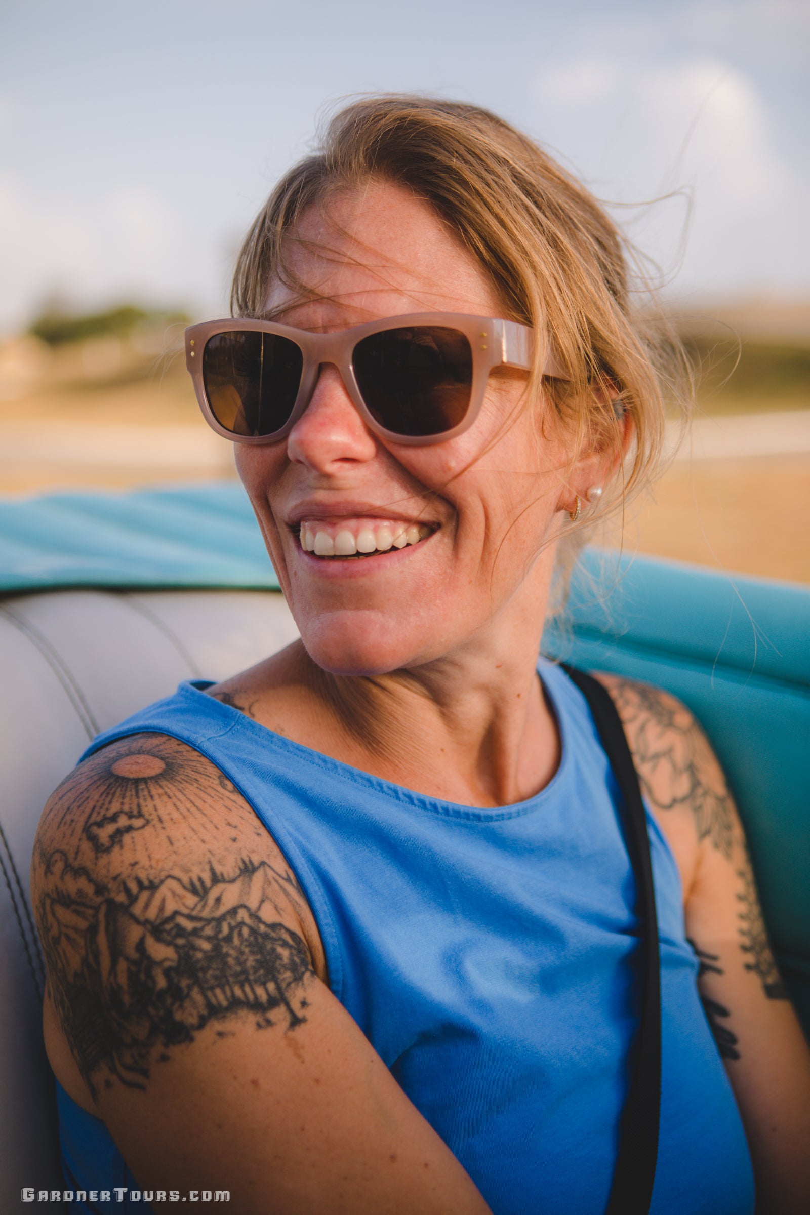 Woman wearing sunglasses and a blue top with tattoos on her arms, sitting in a vehicle.