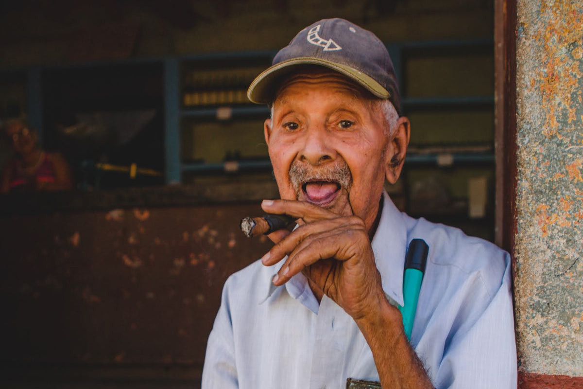A portrait of an elderly Cuban man sitting in a doorway, holding a cigar and smiling, representing the authentic local culture of Cuba.