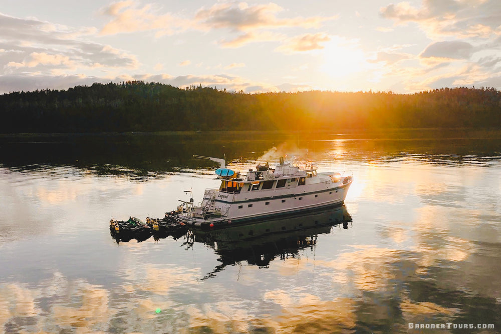 An aerial view of an 84-foot private yacht at anchor in a secluded Alaskan cove during a vibrant golden hour sunset.