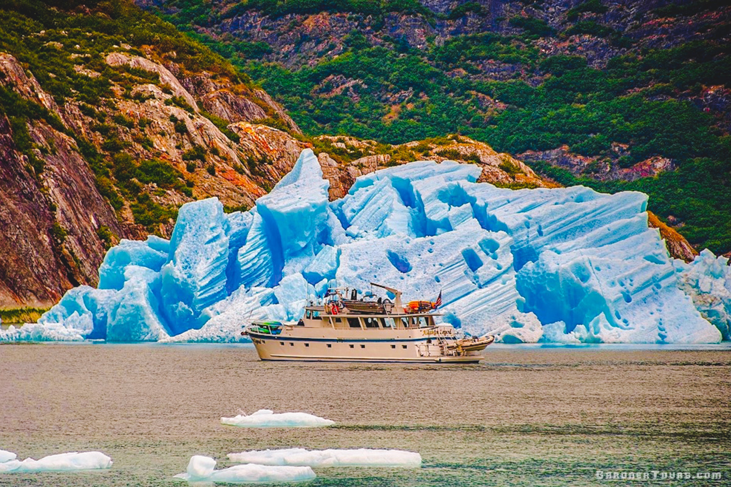 The private yacht navigating safely near a massive, glowing blue glacier wall in Alaska.