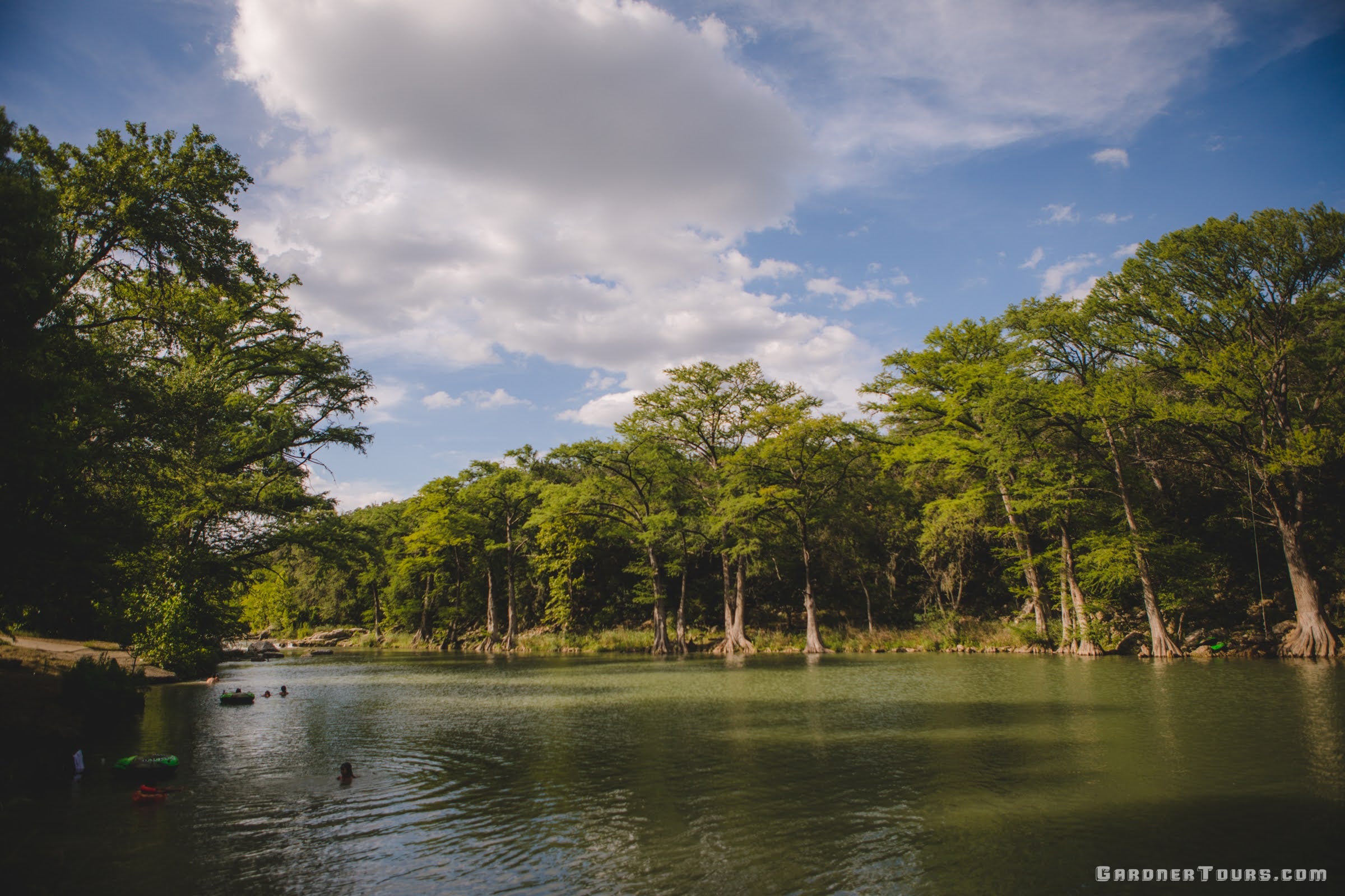 A small group of people relax in the green waters of the Guadalupe River with cypress trees lining the river near New Braunfels, Texas.