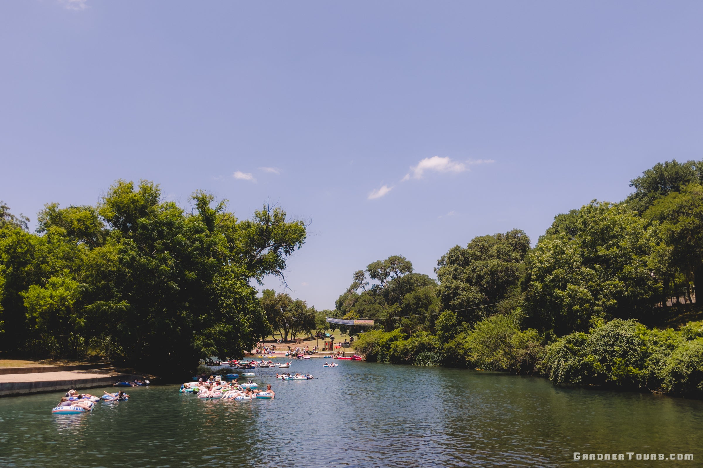Group of people enjoying a day on the Comal River in New Braunfels, Texas with trees and clear sky in the background.
