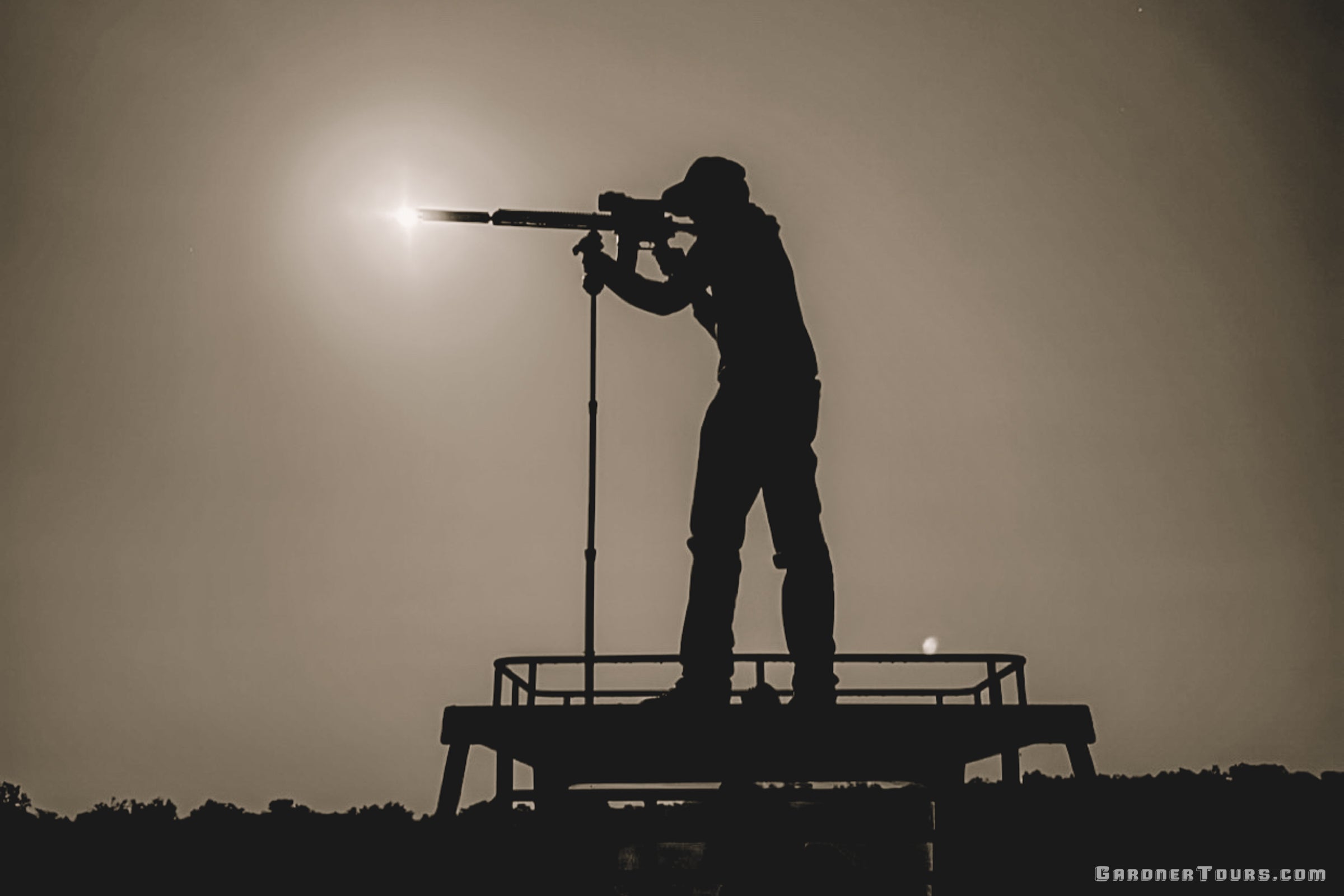 A silhouetted hunter stands on a platform aiming a long gun with a scope, looking for wild hogs during a night thermal hunt in Texas. He is photographed against a bright sun and hazy sky. The image is mostly black silhouette with a warm beige-gray background.