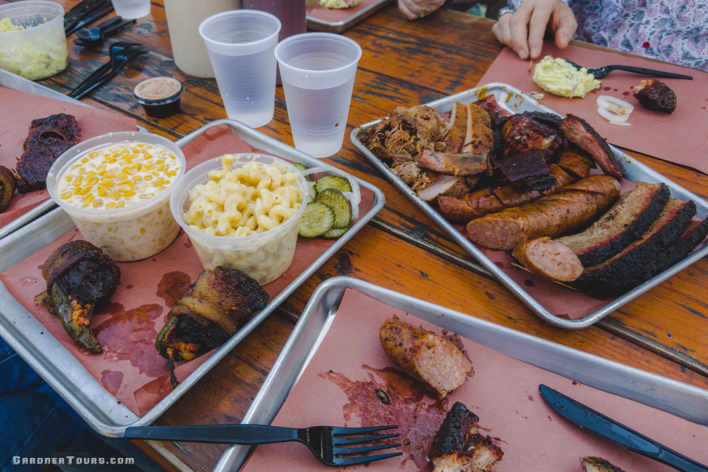 Outdoor setting on a wooden table with a large platter of authentic Texas BBQ with brisket, sausage, turkey, and elotes served during the Gardner Tours True Texas Tour.