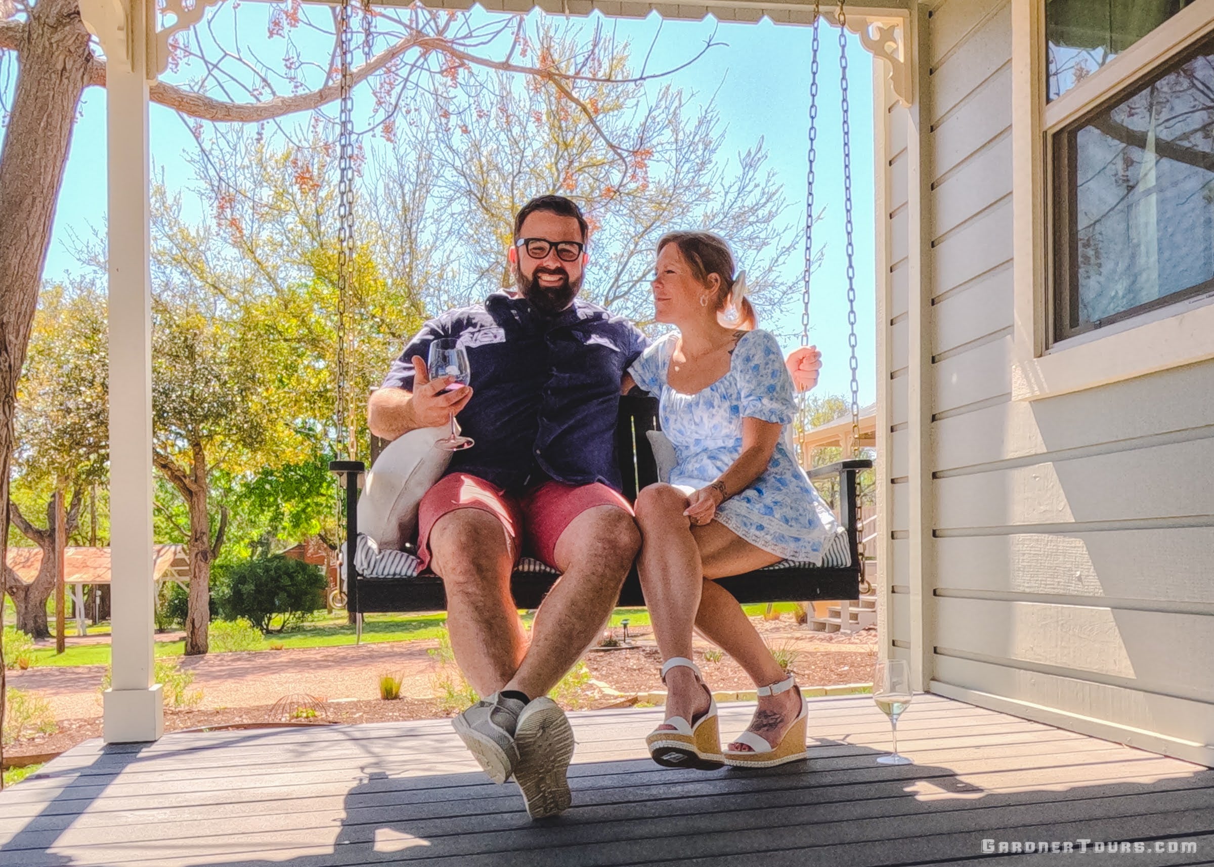 Man and woman sitting on a swing on a porch with trees and a house in the background