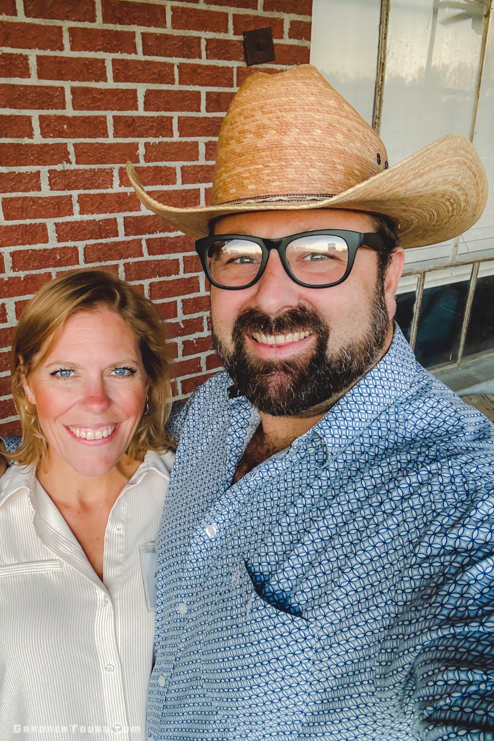 Man wearing a straw cowboy hat and glasses standing next to a woman in front of a brick wall.