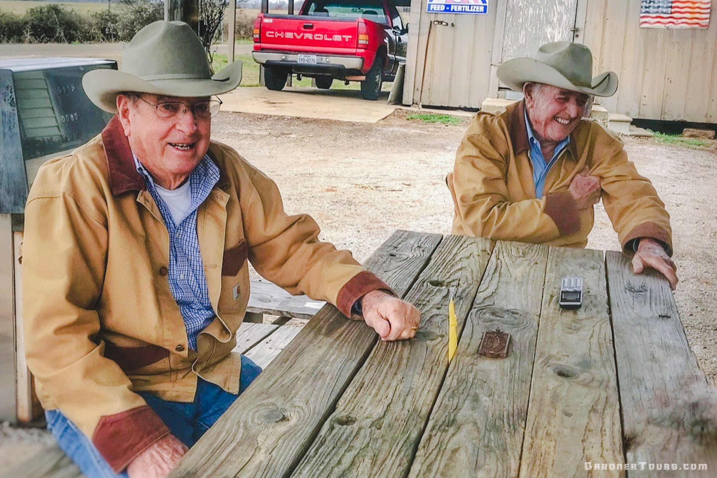 Two men in cowboy hats sitting at a wooden table outdoors with a red truck in the background.
