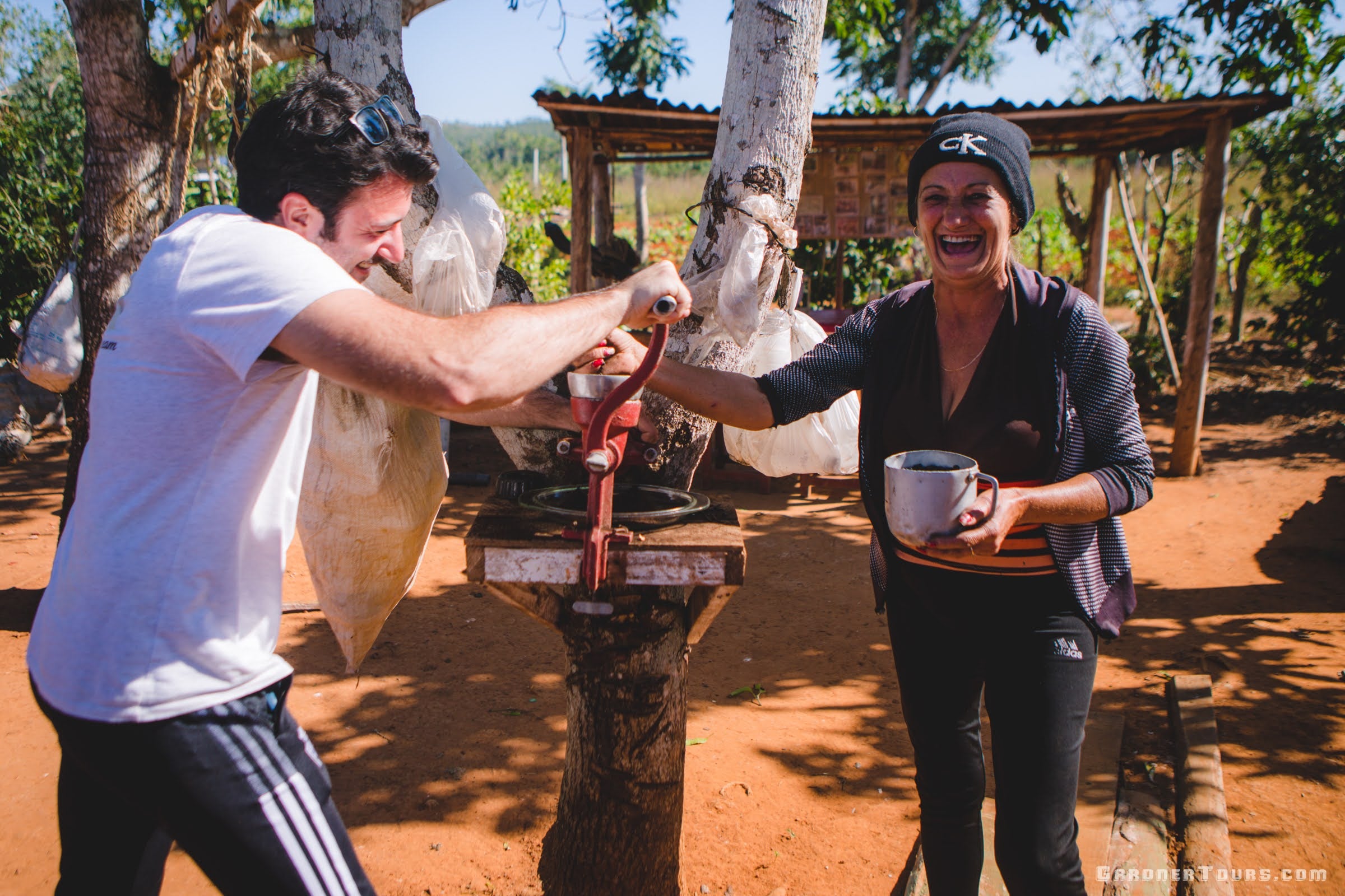 a man tourist grinds coffee by hand and a local woman laughs at him on the Coffee Farm in Vinales Cuba