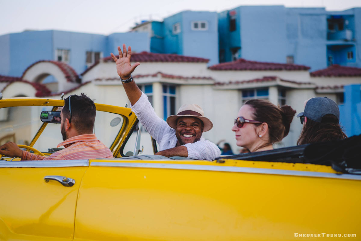 Gardner Tours Cuban Tour Guide Michel waving and smiling as he rides in a yellow classic car through Havana, Cuba with his American travelers