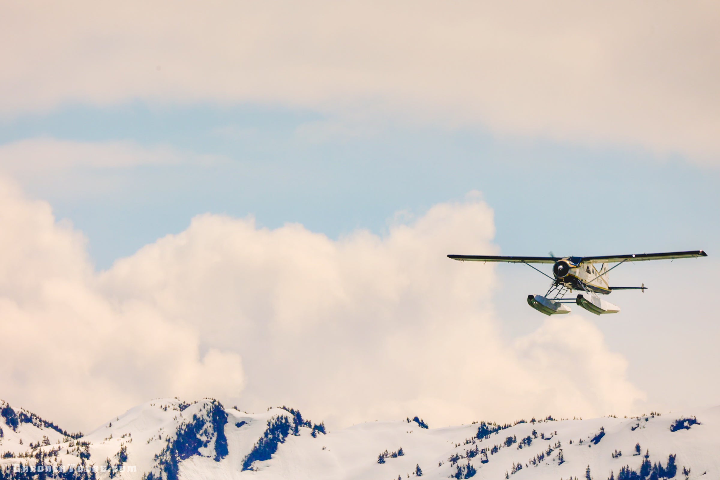 An Alaskan floatplane soaring over snow-capped mountains, representing the arrival and travel preparation for a private yacht journey in Southeast Alaska.