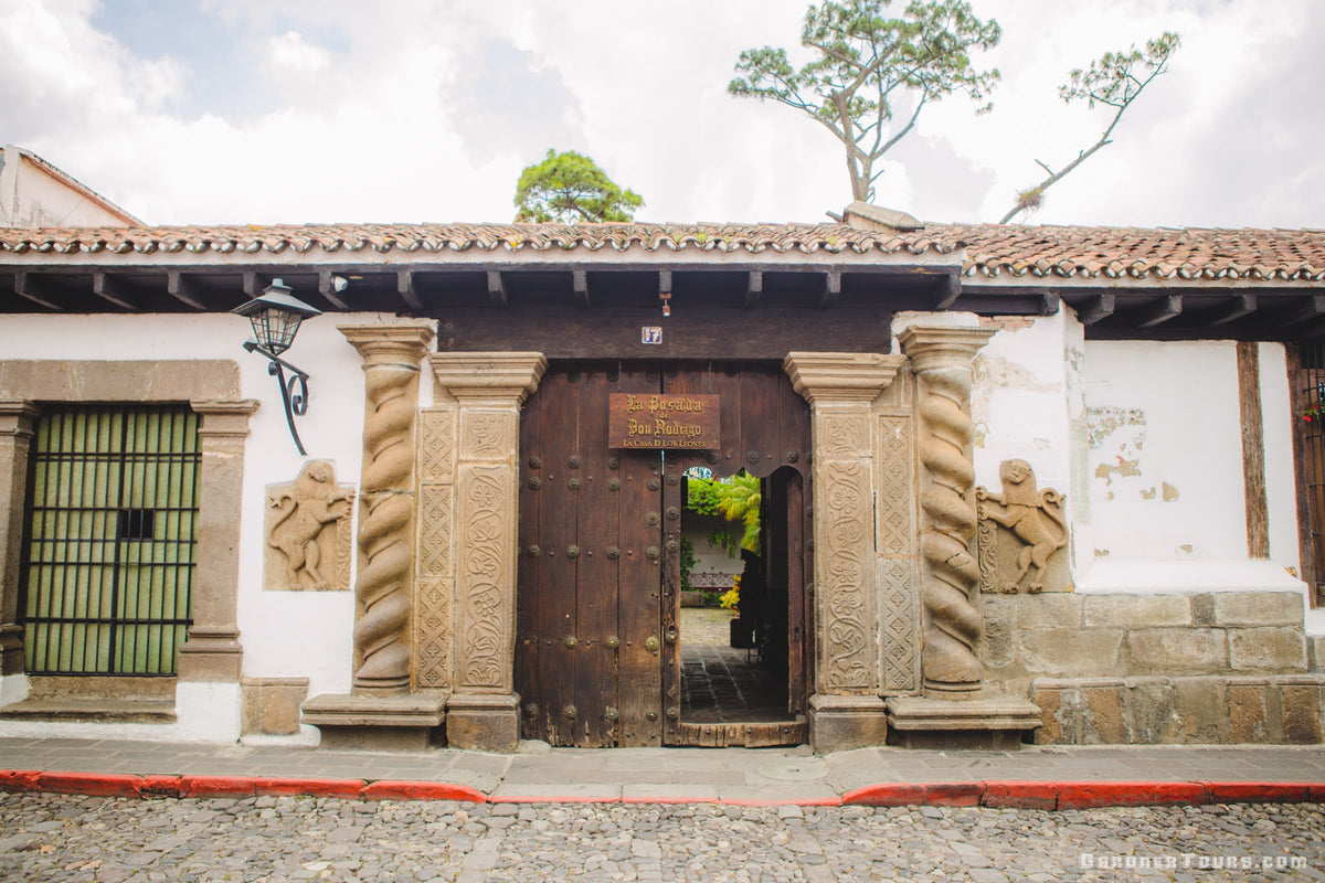 The entrance to a historic colonial-style building featuring a large dark wooden double door, carved stone pillars, a tiled roof, and a cobblestone street in Antigua, Guatemala.