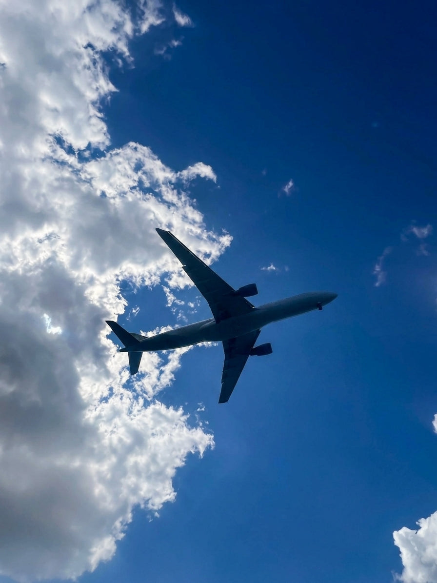 A dramatic low-angle, sharp photograph looking up at the precise silhouette of a large, twin-engine commercial airliner. The plane is perfectly centered, silhouetted against a deep blue sky with complex, sun-dappled and brilliantly backlit cumulus and altocumulus clouds. All lines of the aircraft and textures of the clouds are in sharp focus.