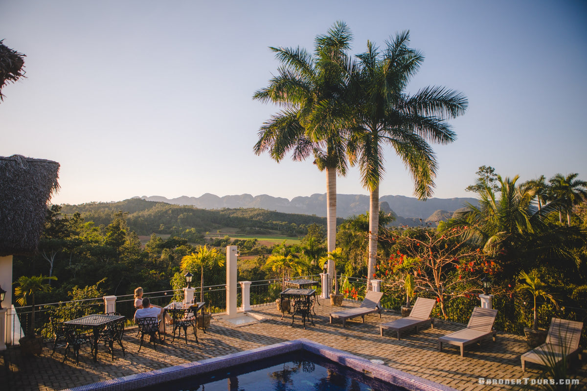 Travelers relaxing on a sunlit patio deck surrounded by palm trees overlooking Viñales Valley in Cuba