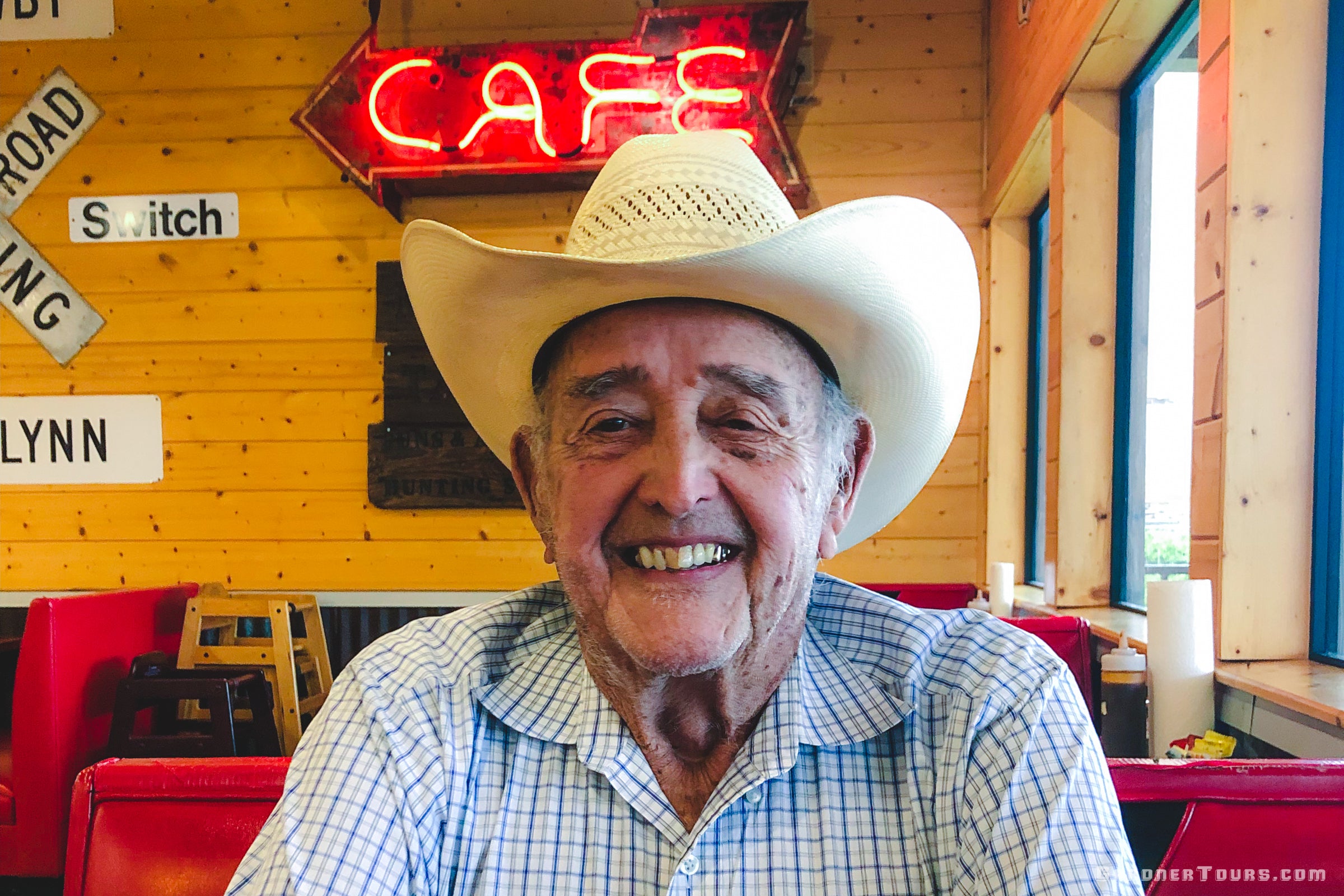 Close-up of Homer Coleman, an elderly gentleman, sitting at a café table with a big café neon sign in the background, enjoying a quiet moment during the Gardner Tours Texas Tours experience.