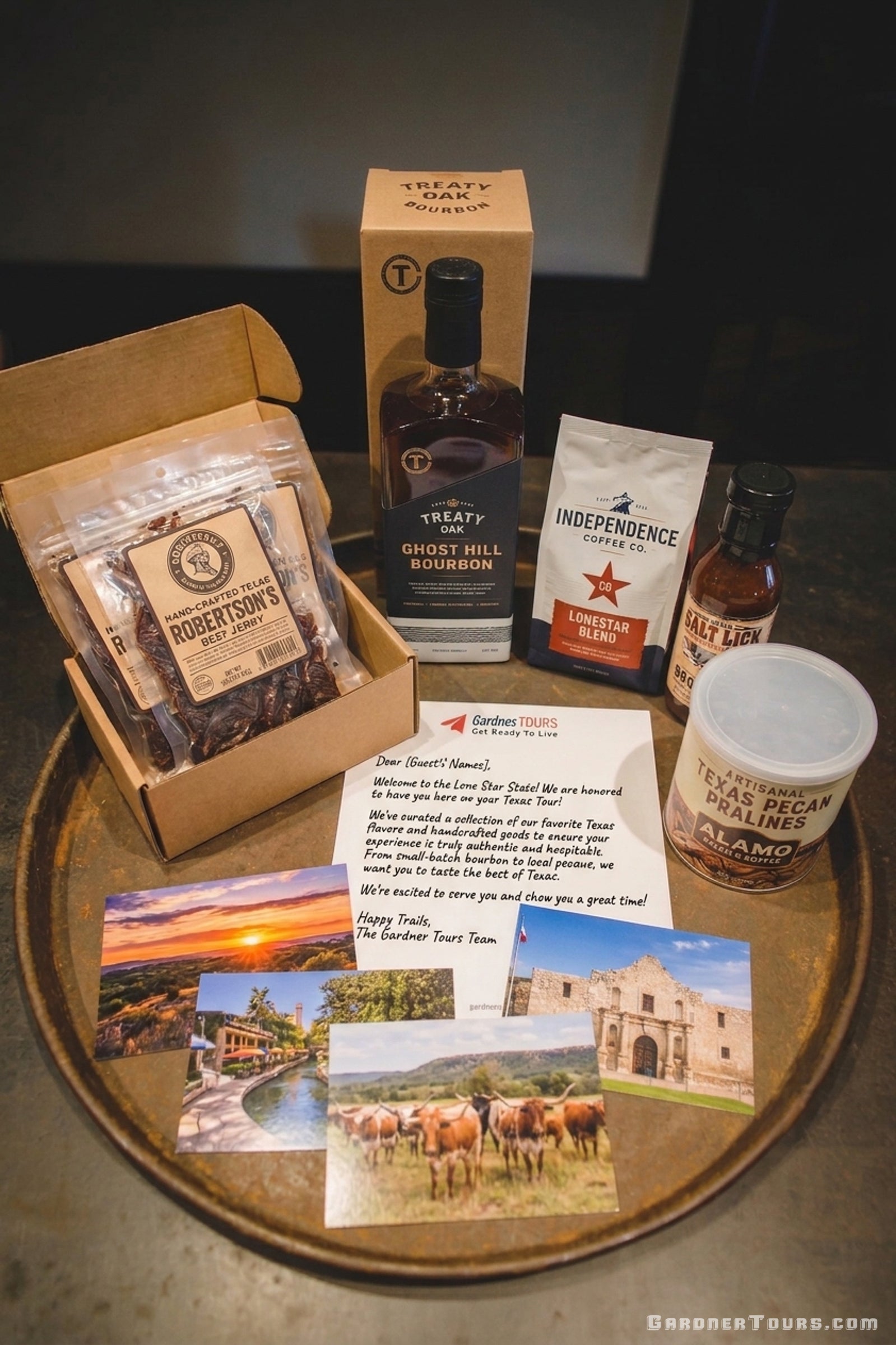 A Gardner Tours Texas Welcome Package displayed on a rustic metal tray, featuring Treaty Oak Ghost Hill Bourbon, Robertson's Beef Jerky, Independence Coffee, Salt Lick BBQ Sauce, Texas Pecan Pralines, a personalized welcome letter, and postcards of Texas scenery and the Alamo.