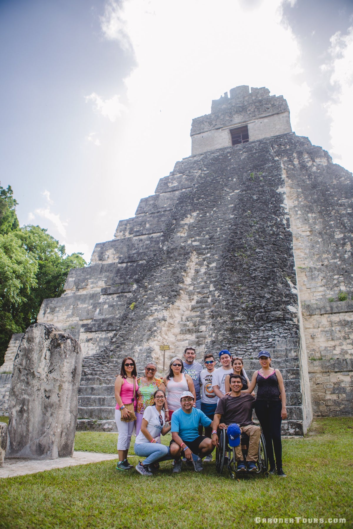 Group of tourists posing in front of a large stone Mayan Pyramid temple with greenery around.