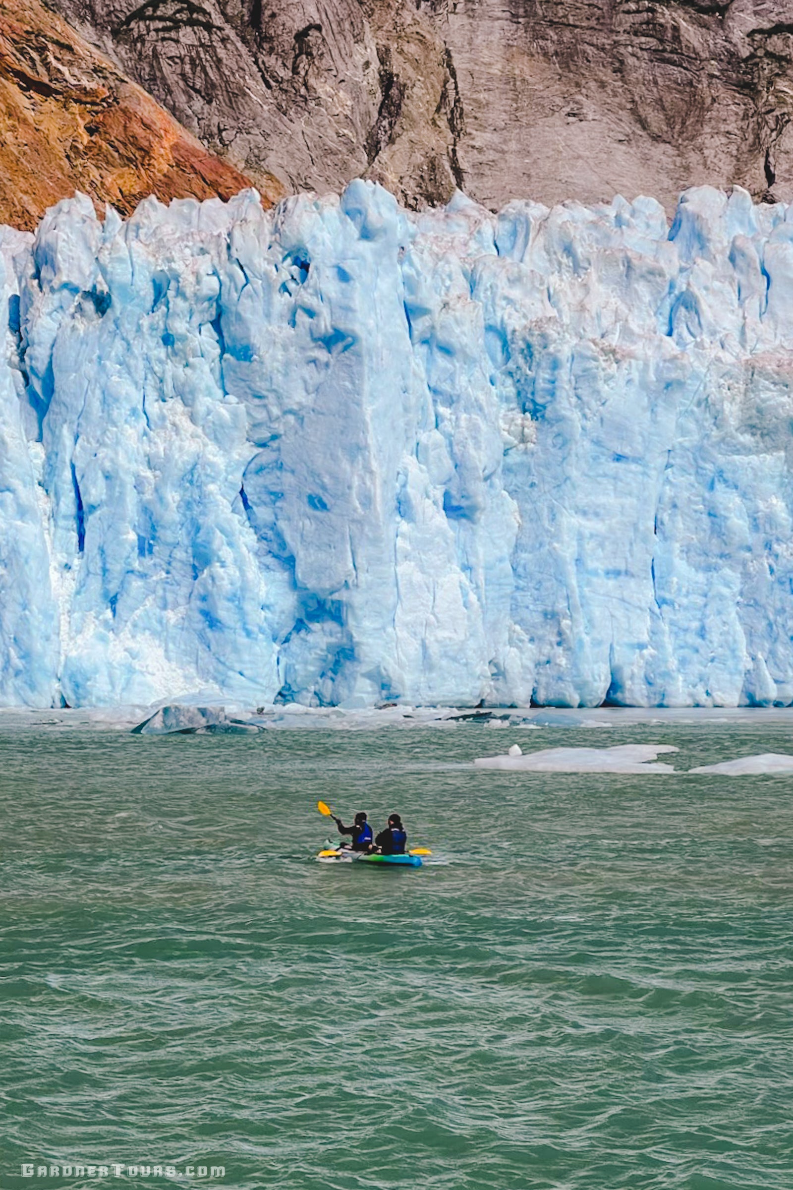 Two travelers in a tandem kayak paddling in front of a towering blue tidewater glacier wall in Alaska.