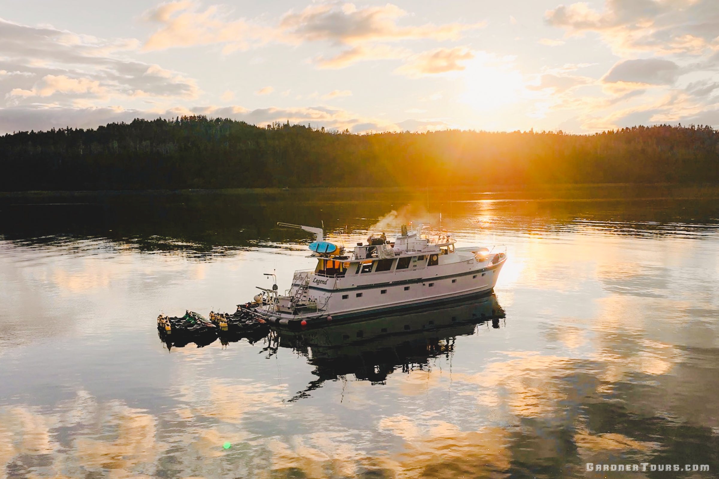An aerial view of an 84-foot private yacht at anchor in a secluded Alaskan cove during a vibrant golden hour sunset.