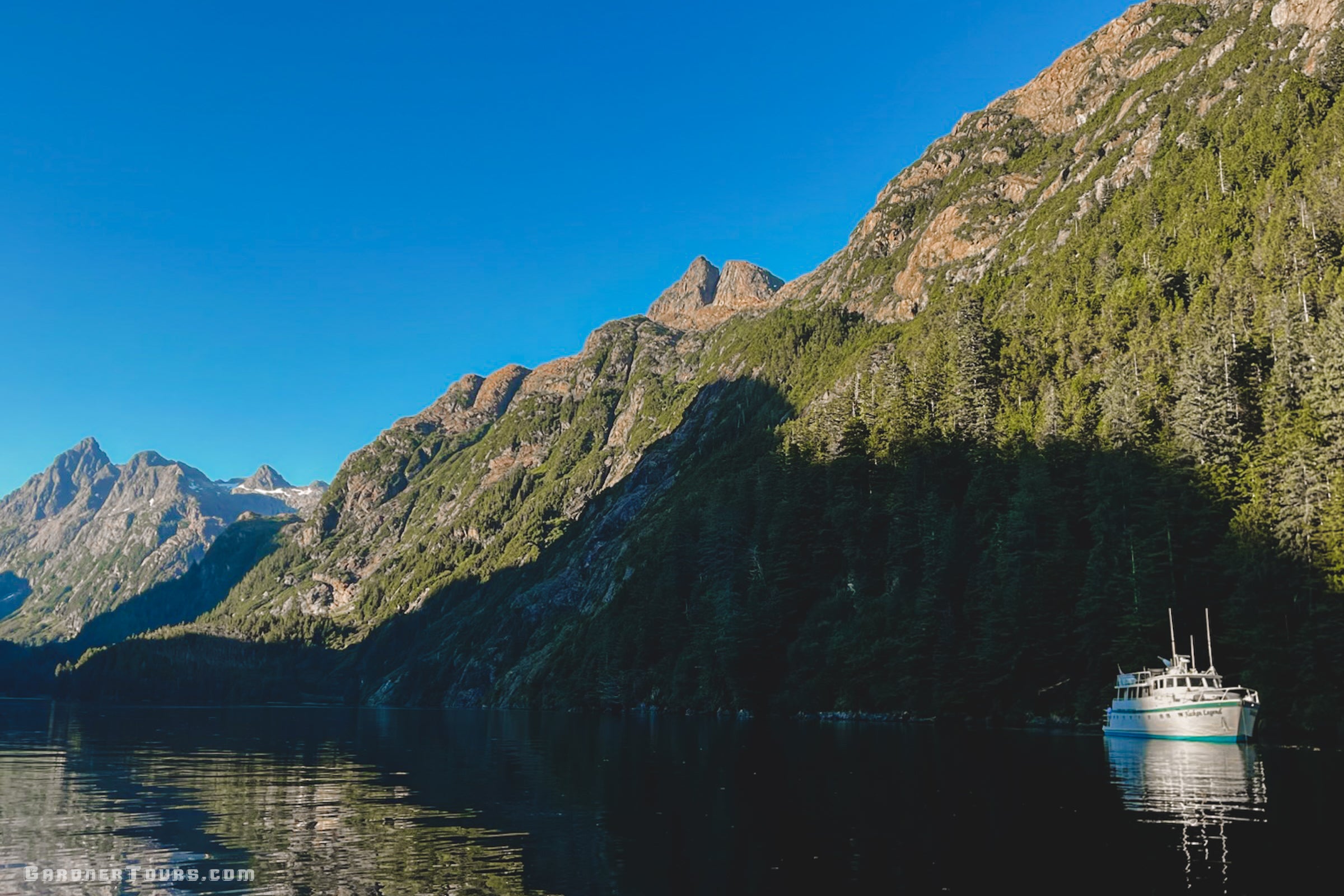 A private 84-foot luxury yacht anchored beneath towering granite cliffs and lush evergreen forests in a secluded Alaska fjord during an Inside Passage expedition.