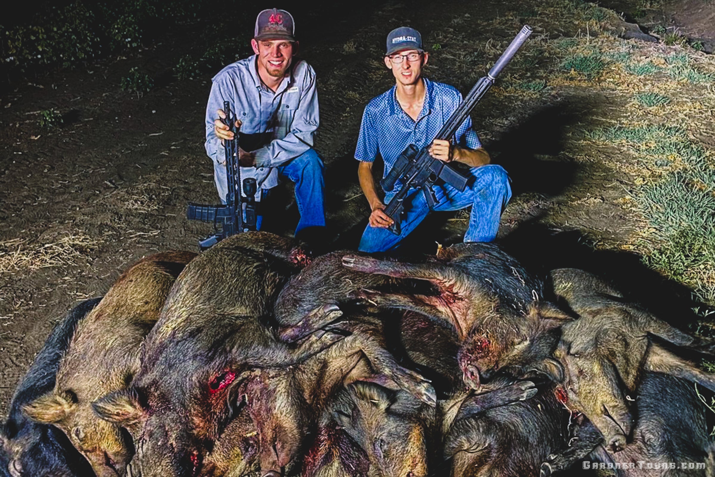 Two happy hunters kneeling behind a pile of harvested wild hogs with their thermal rifles after a successful night hunt in Texas.