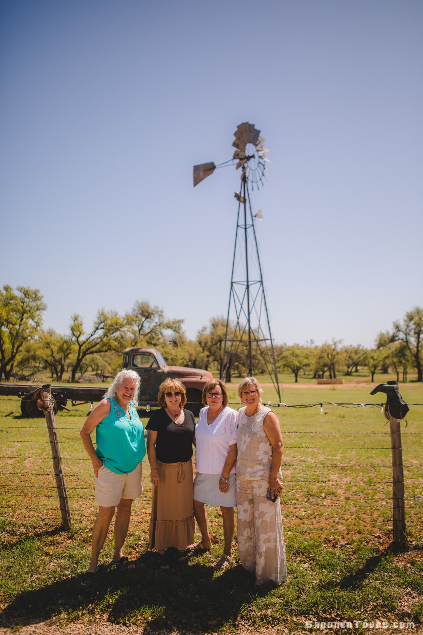 A group of four middle aged women pose in front of a barbed wire fence with a rustic classic Chevrolet pickup and a windmill on the willow city loop outside of Fredericksburg, Texas.
