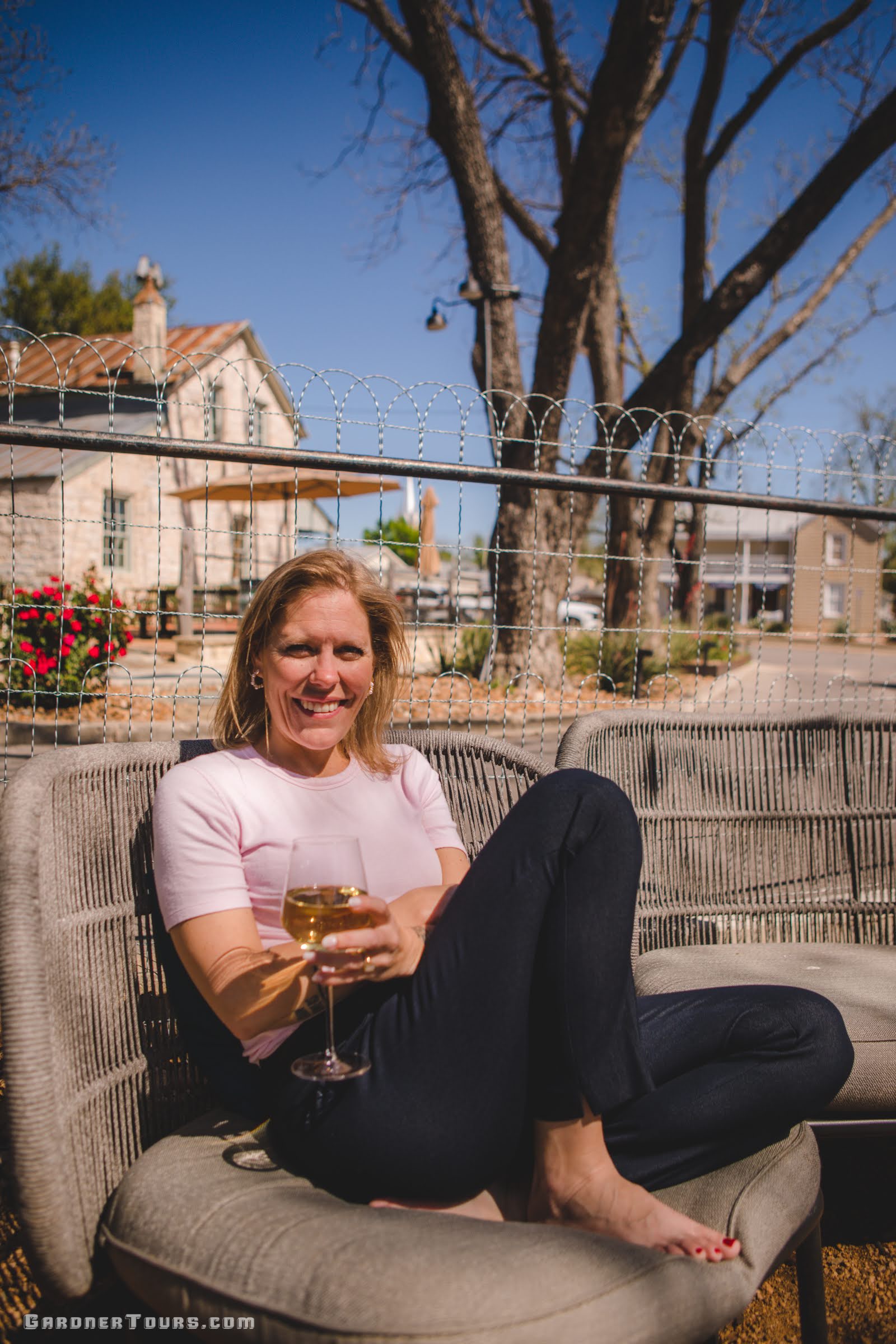 Young woman, Josie Gardner, enjoys a glass of white wine, while sitting on a tan chair under the oak trees outside of Hill and Vine Restaurant in Fredericksburg, Texas.