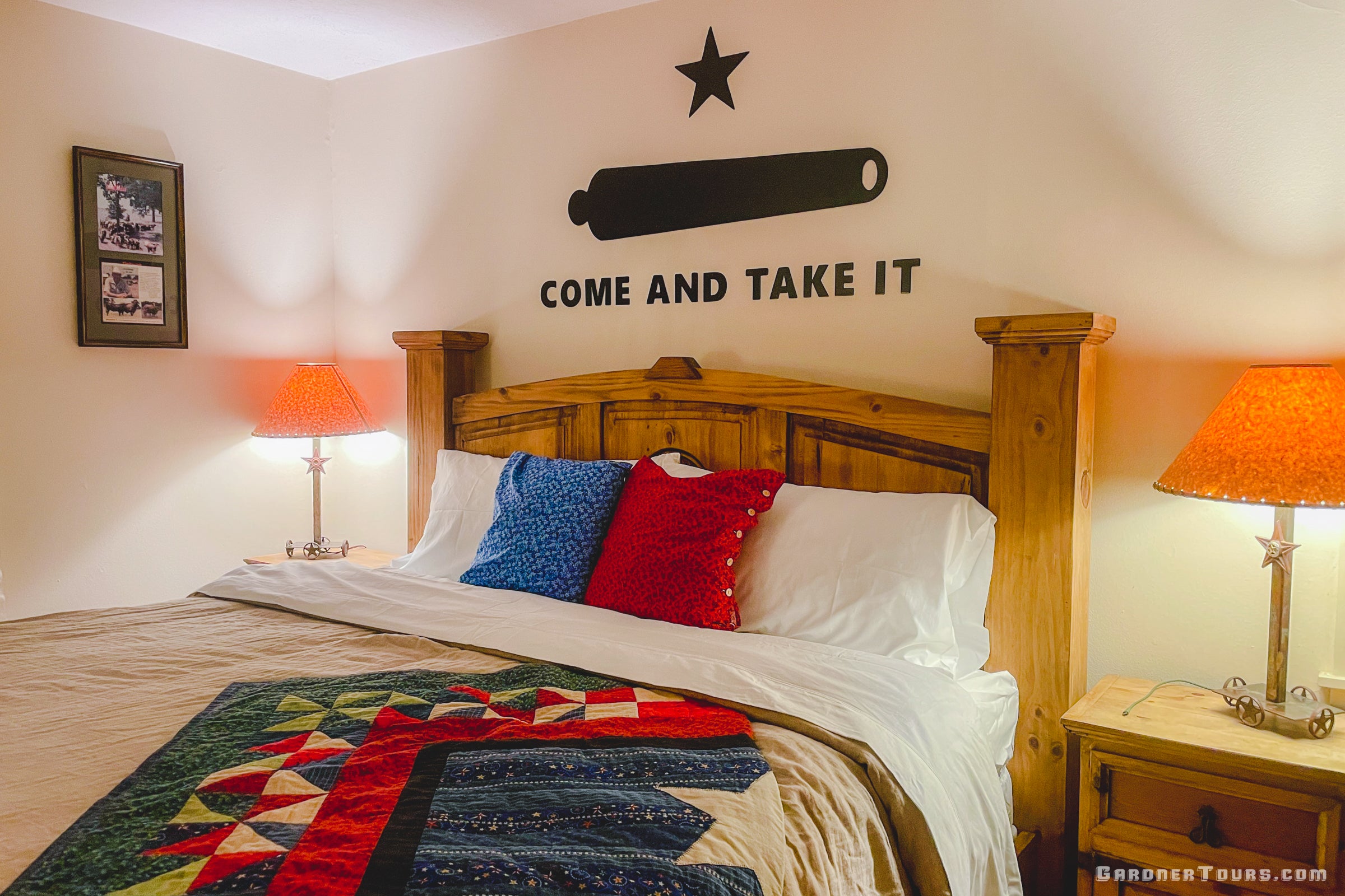 A Texas-themed room with a rustic wooden bed in a cozy BnB, featuring a colorful Texas quilt, linen bedding, two red and blue decorative pillows, and a black 'COME AND TAKE IT' cannon graphic mounted on the wall above the headboard.