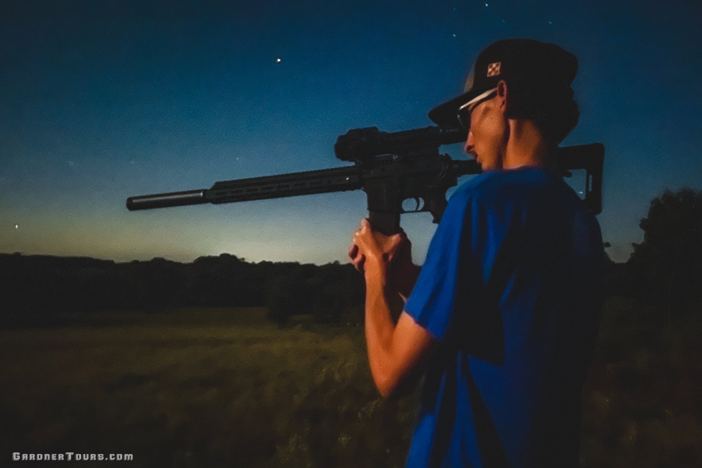 Hunter aiming a thermal rifle with a suppressor against a dusk sky during a Texas thermal night hog hunt.