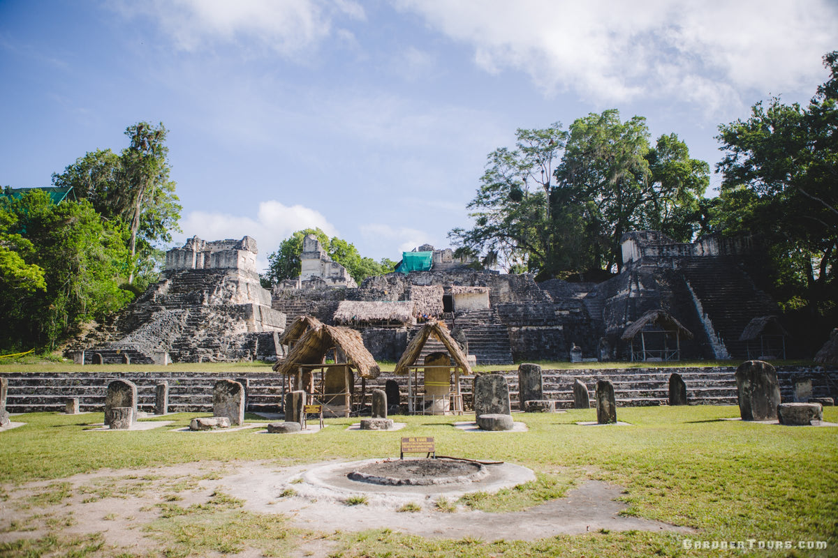 Mayan ruins at Tikal with stone structures, thatched-roof huts, grassy fields, and trees under a blue sky with clouds.